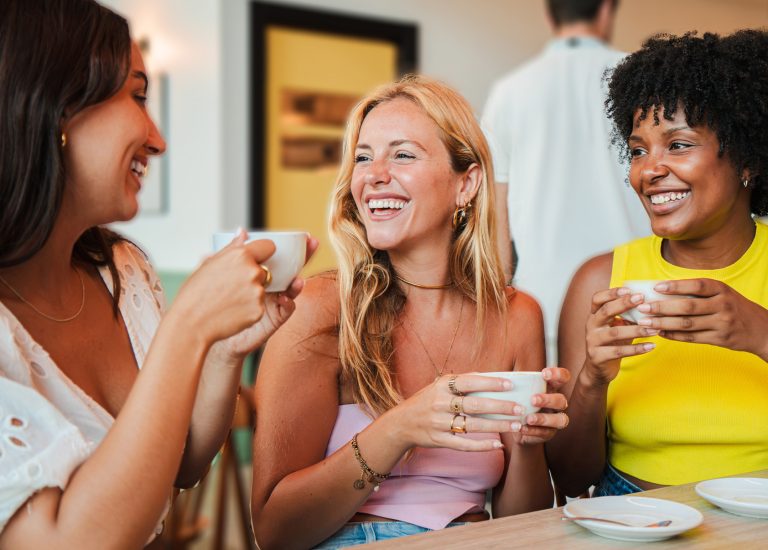 Three friends sitting at a café table laughing and drinking coffee together