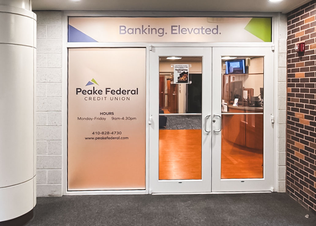 Entrance to Peake Federal Credit Union, featuring a frosted window with the credit union’s logo, business hours, and contact information, alongside glass doors showing the interior lobby. A banner above reads ‘Banking. Elevated.