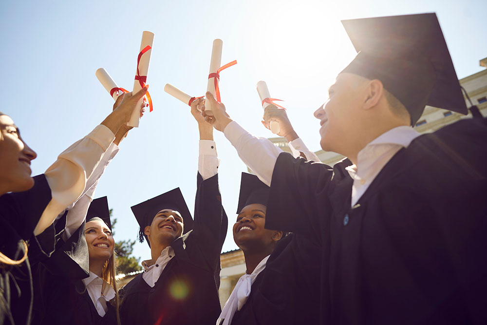 Graduates in caps and gowns raising their diplomas together in celebration under bright sunlight.