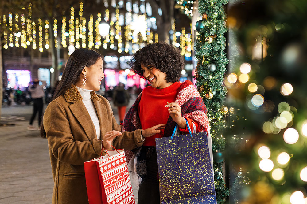 How Friends Can Help Protect Wallets. Two friends holiday shopping together, smiling and comparing purchases while keeping each other accountable on seasonal spending.