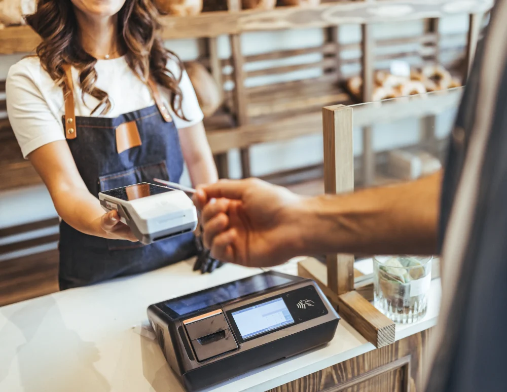 Smiling Cashier Hands Card Reader to Customer at Bakery Checkout in Busy Store