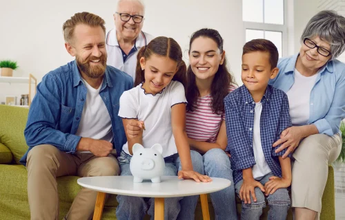 Girl is putting a coin in family white piggy bank while all family is looking at her at home. Smiling grandparents, parents and kids are saving money together.