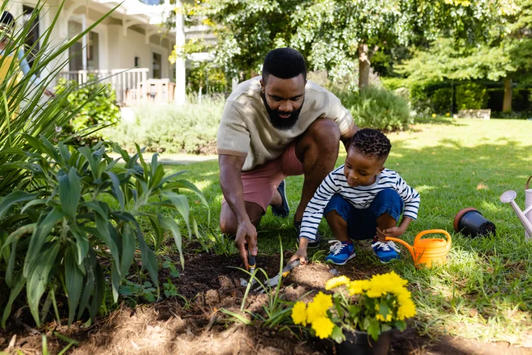 African american father and son digging dirt with tools on grassy field in backyard